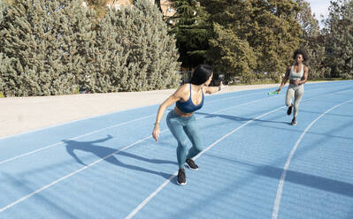 Fit multirassische weibliche Athleten, die den Staffelstab weitergeben, während sie während eines Staffellaufs im Stadion entlang der Bahn laufen - ADSF22521