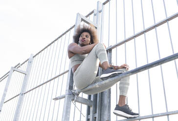 Eine afroamerikanische Sportlerin in Sportkleidung sitzt während des Trainings auf einem Sportplatz auf einem Reck aus Metall und schaut weg - ADSF22523