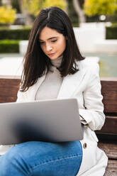 Serious female entrepreneur sitting on bench in urban park and typing on laptop while working remotely on business project - ADSF22532