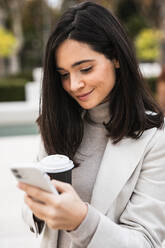 Lächelnde Unternehmerin, die auf der Straße ihr Smartphone benutzt und Kaffee trinkt - ADSF22534