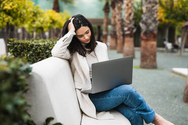 Nachdenkliche Unternehmerin mit Laptop, die auf einer Bank im Stadtpark sitzt und online an einem Projekt arbeitet - ADSF22535