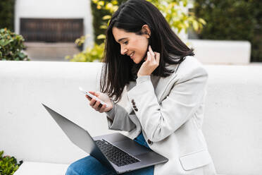 Glückliche Geschäftsfrau, die mit ihrem Laptop auf einer Bank sitzt und auf ihrem Smartphone surft, während sie im Stadtpark arbeitet - ADSF22537