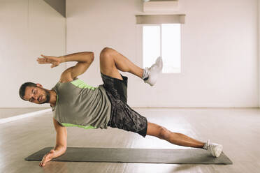 Full body of young focused fit determined sportsman in activewear performing High Side Plank with leg raises exercise during workout in studio near big wall mirror - ADSF22542