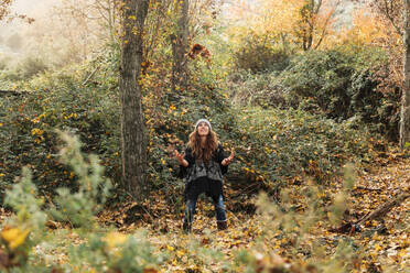 Verspielte Frau wirft Herbstblätter, während sie im Wald steht - MRRF00998