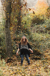 Frau betrachtet Herbstblätter in der Luft, während sie in einem Wald im Urlaub steht - MRRF00999