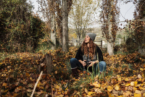 Woman with camera looking away while sitting amidst autumn leaves in forest - MRRF01002