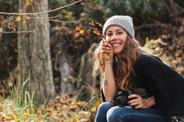Lächelnde schöne Frau, die Blätter hält, während sie mit der Kamera im Wald im Herbst sitzt - MRRF01006