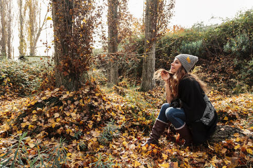 Lächelnde Frau mit Hand im Haar, die im Herbstwald sitzt und nachdenkt - MRRF01007