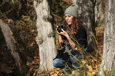 Thoughtful woman with camera looking away while crouching amidst trees in forest during vacations - MRRF01009