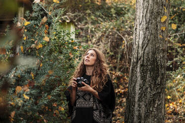 Mittlere erwachsene Frau schaut nach oben, während sie die Kamera an einem Baumstamm im Wald hält - MRRF01013