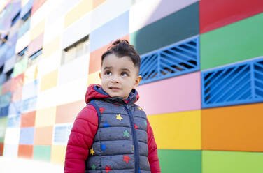 Boy in padded jacket contemplating while standing by colorful wall - JCCMF01802