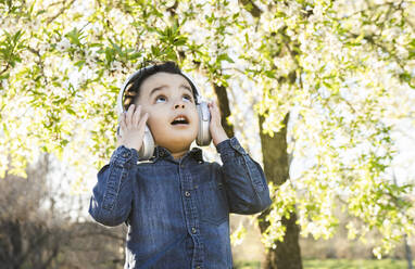 Überraschender Junge, der aufschaut, während er im Park Musik über drahtlose Kopfhörer hört - JCCMF01814