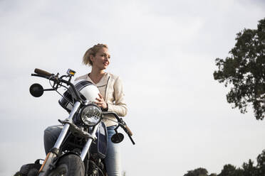 Beautiful female biker holding helmet on motorcycle while looking away in front of sky - ABZF03519