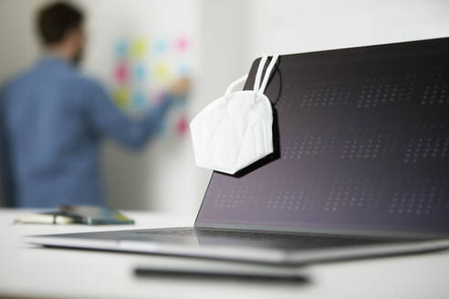 Face mask hanging on laptop while man standing in background at office - PSBHF00014
