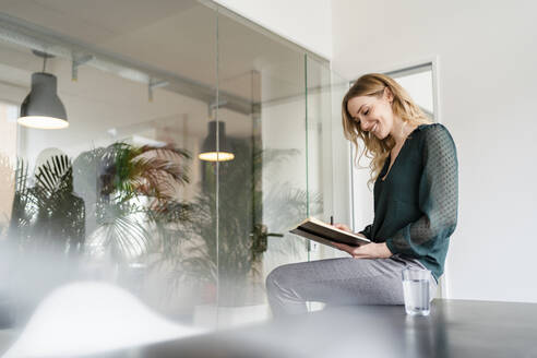 Lächelnde Unternehmerin liest Tagebuch, während sie auf einem Konferenztisch im Büro sitzt - DIGF15009