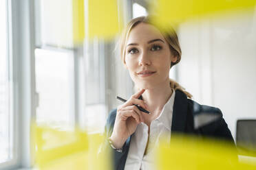 Thoughtful businesswoman with pen looking at adhesive note seen through glass - DIGF15025