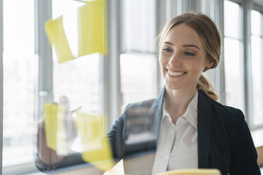 Lächelnde Geschäftsfrau, die auf Haftnotizen schreibt, gesehen durch Glas im Büro - DIGF15030