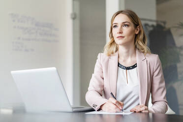 Businesswoman contemplating while writing on document by laptop in office - DIGF15046