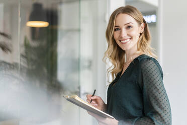 Smiling businesswoman holding diary and pen at office - DIGF15070