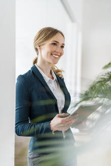 Lächelnde Geschäftsfrau, die wegschaut und ein digitales Tablet im Büro hält - DIGF15082