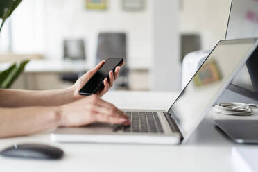 Female professional holding mobile phone while working on laptop in office - DIGF15104