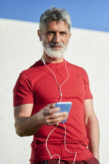 Älterer Mann mit Kopfhörern und Smartphone an der Wand - KIJF03700