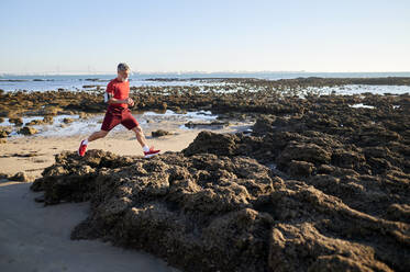 Männlicher Jogger läuft auf Felsen am Strand - KIJF03716