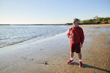 Man holding water bottle standing on seashore at beach during sunset - KIJF03720