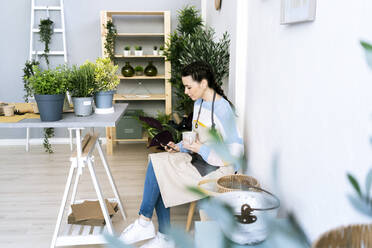 Frau mit Tasse, die in der Werkstatt sitzend ihr Smartphone benutzt - GIOF12096