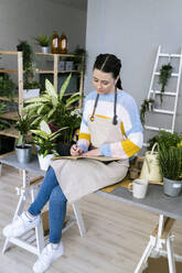 Junge Frau schreibt in ein Buch, während sie auf einem Tisch in einer Werkstatt sitzt - GIOF12101