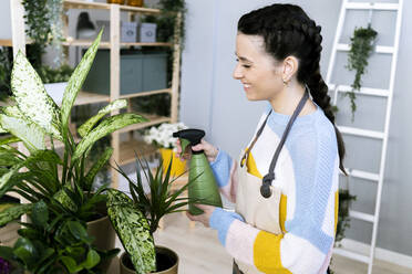 Smiling woman spraying water on plants while working at workshop - GIOF12104
