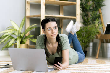 Contemplating woman holding smart phone by laptop while lying at home - GIOF12118