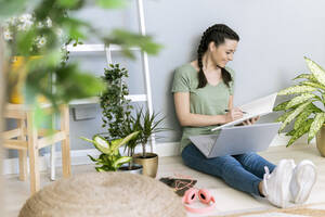 Junge Frau beim Schreiben in einem Buch, während sie zu Hause an der Wand sitzt - GIOF12128