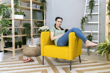 Junge Frau entspannt sich auf der Couch inmitten von Pflanzen zu Hause - GIOF12130