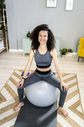 Glückliche Frau sitzt auf einem Fitnessball im Wohnzimmer - GIOF12156