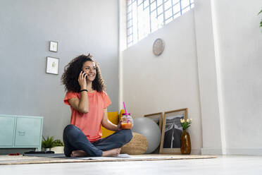 Lächelnde Frau, die mit einem Mobiltelefon spricht, während sie zu Hause ein Saftglas in der Hand hält - GIOF12162