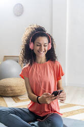 Glückliche Frau, die ein Smartphone benutzt, während sie zu Hause über Kopfhörer Musik hört - GIOF12171