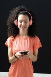 Lächelnde Frau, die Musik hört, während sie ein Mobiltelefon vor einer Wand benutzt - GIOF12173