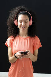 Smiling woman listening music while using mobile phone in front of wall - GIOF12173