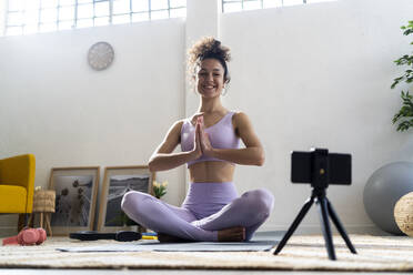 Smiling female influencer with hands clasped vlogging while doing yoga at home - GIOF12178