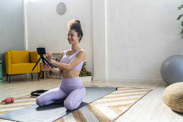 Smiling woman kneeling while touching mobile phone during vlogging at home - GIOF12184