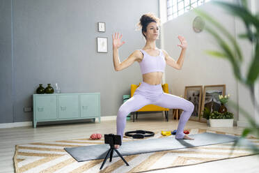Young woman doing yoga while vlogging through smart phone in living room - GIOF12188
