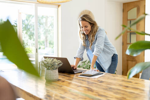 Lächelnde Geschäftsfrau, die einen Laptop benutzt und sich zu Hause auf einen Tisch stützt - SBOF03699