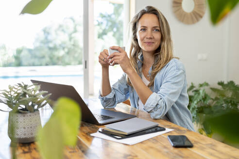 Weibliche Unternehmerin sitzt mit Laptop und Kaffeetasse und schaut weg - SBOF03705