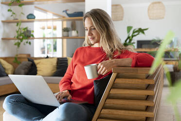 Blonde Frau benutzt einen Laptop und hält eine Kaffeetasse im Wohnzimmer - SBOF03713