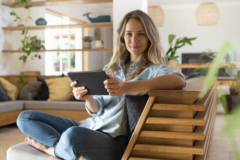 Glückliche Frau, die ein Tablet hält und mit gekreuzten Beinen auf der Couch im Wohnzimmer sitzt - SBOF03718