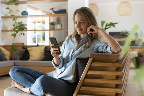 Blondhaarige Frau, die im Wohnzimmer sitzend eine SMS über ihr Smartphone verschickt - SBOF03719