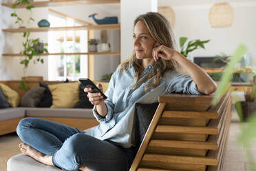 Smiling woman sitting with mobile phone on couch while looking away - SBOF03720