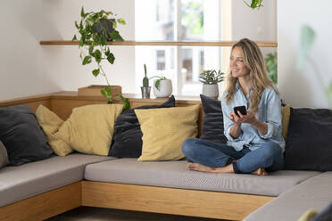Lächelnde blonde Frau, die ihr Smartphone in der Hand hält, während sie zu Hause auf der Couch im Wohnzimmer sitzt - SBOF03721
