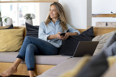 Woman sitting with mobile phone and laptop on couch n living room - SBOF03723